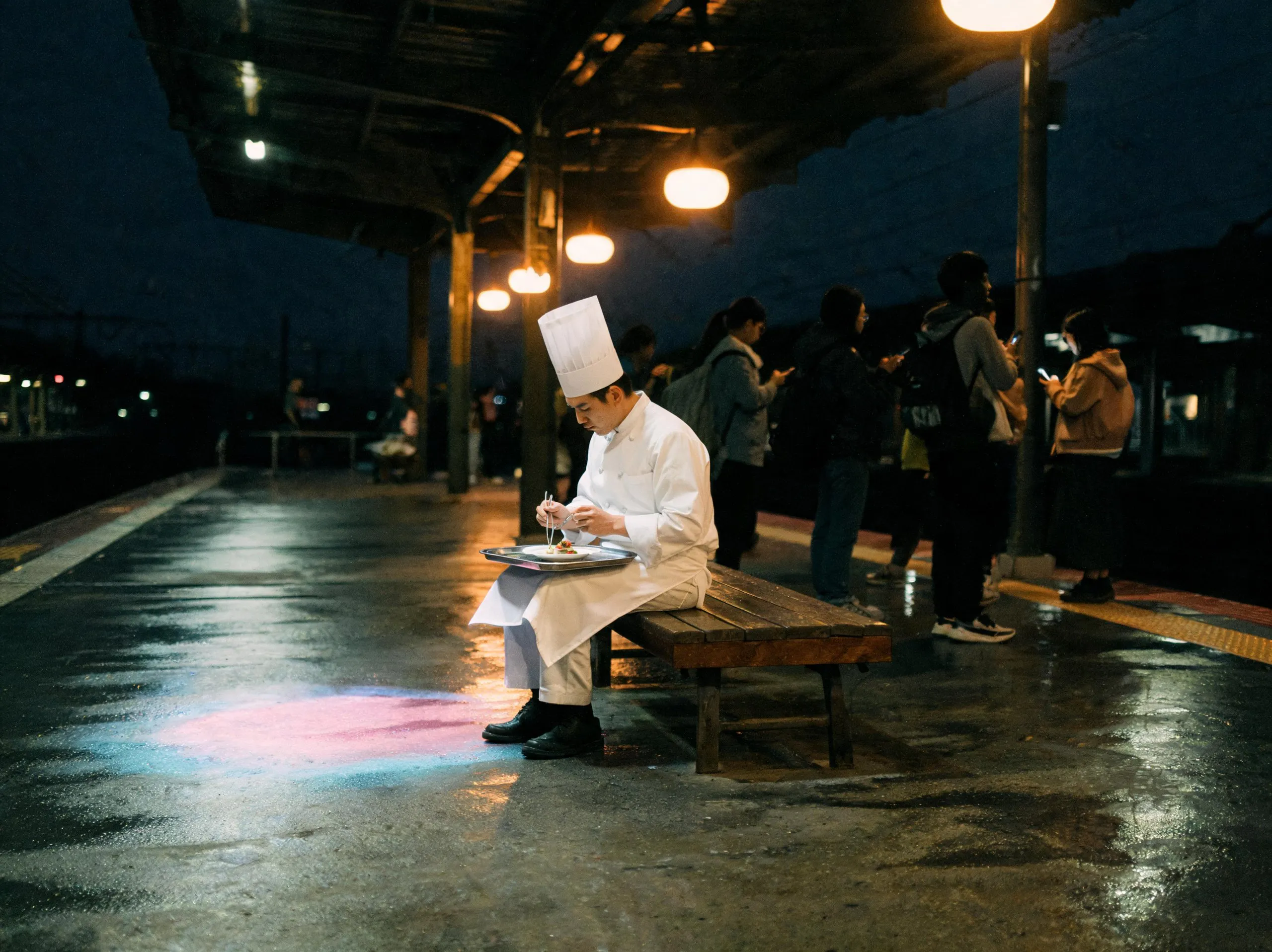 A chef in whites eating a plated meal on a rainy train platform at night as commuters wait